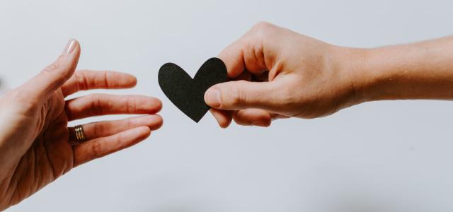 Image of two hands against a white background. One of the hands is passing a black paper heart to the other one